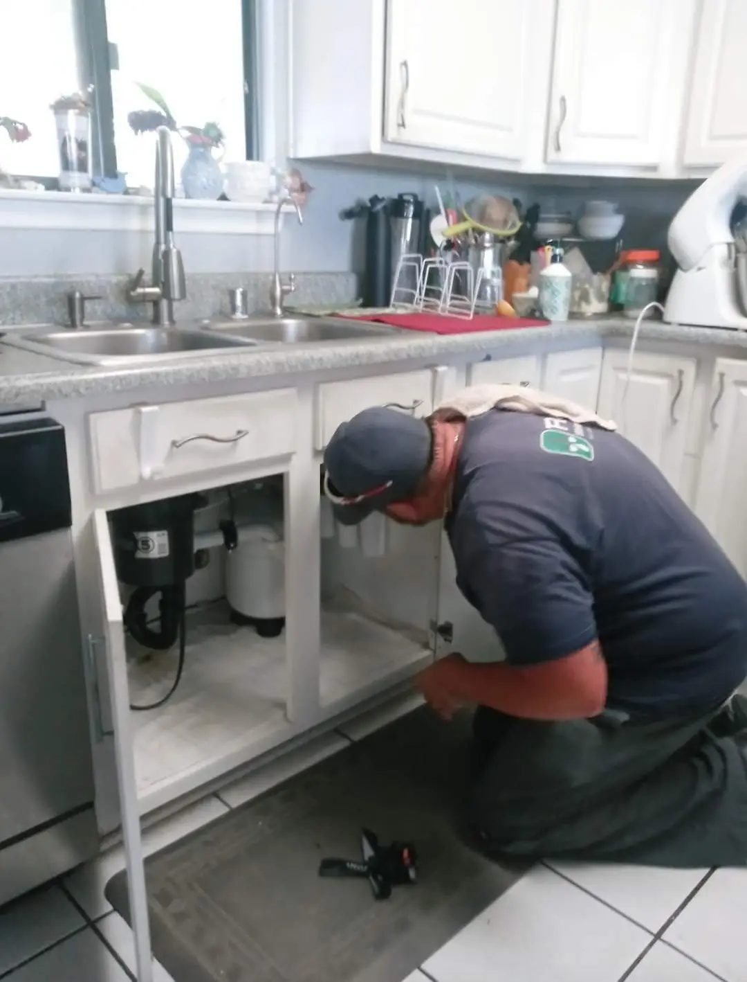 A technician crouches beside a kitchen sink, inspecting pipes under the cabinet while tools are laid out on the floor.